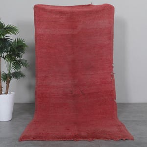 May include: A solid red rug with a slightly distressed look. The rug is folded in half and standing upright on a gray floor.