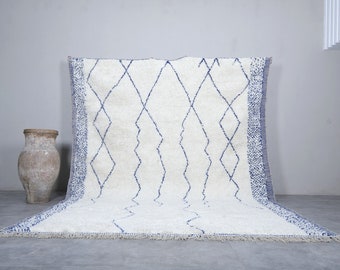 May include: A white and blue Moroccan rug with a diamond pattern. The rug has a fringe border and is made of wool.