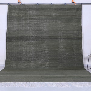 May include: A dark green rug with a subtle pattern and white fringe. The rug is hanging on a wall with a chair in the foreground.