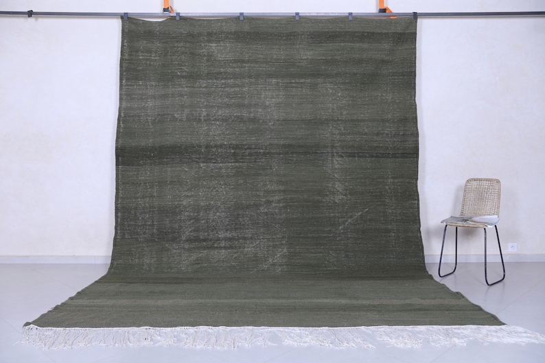 May include: A dark green rug with a subtle pattern and white fringe. The rug is hanging on a wall with a chair in the foreground.