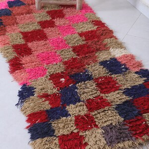 May include: A colourful, hand-woven rug with a checkered pattern in shades of pink, red, beige, and blue. The rug has a shaggy texture and is made with natural fibres.