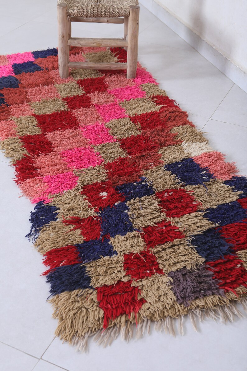 May include: A colourful, hand-woven rug with a checkered pattern in shades of pink, red, beige, and blue. The rug has a shaggy texture and is made with natural fibres.