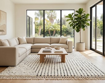 May include: A modern living room with a large beige sectional sofa, a wooden coffee table, and a patterned rug. A large potted plant sits near a window overlooking a lush outdoor area. The room has a neutral color palette with natural light.
