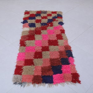 May include: A long, rectangular rug with a checkered pattern in shades of pink, red, brown, and blue. The rug has a fringed edge.