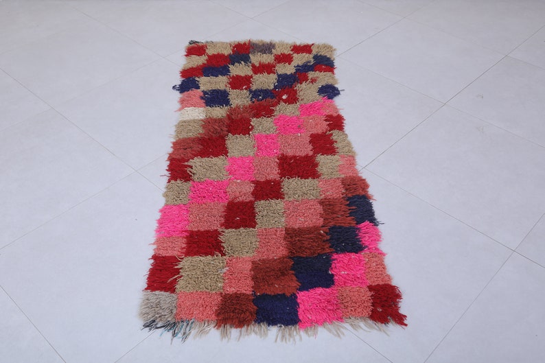 May include: A long, rectangular rug with a checkered pattern in shades of pink, red, brown, and blue. The rug has a fringed edge.