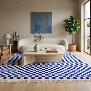 May include: A modern living room featuring a blue and white checkered rug with fringe. A light-colored sofa, wooden coffee table, and a leather armchair are arranged on the rug. A large window and a painting complete the room's decor.