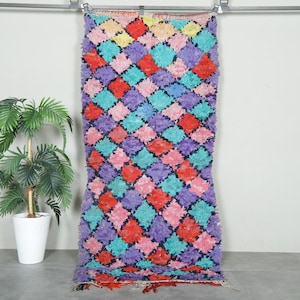 May include: A vibrant rug with a diamond pattern in red, pink, purple, turquoise, and yellow. The rug is suspended against a white wall, with a potted plant to the left. The rug has a shaggy texture.