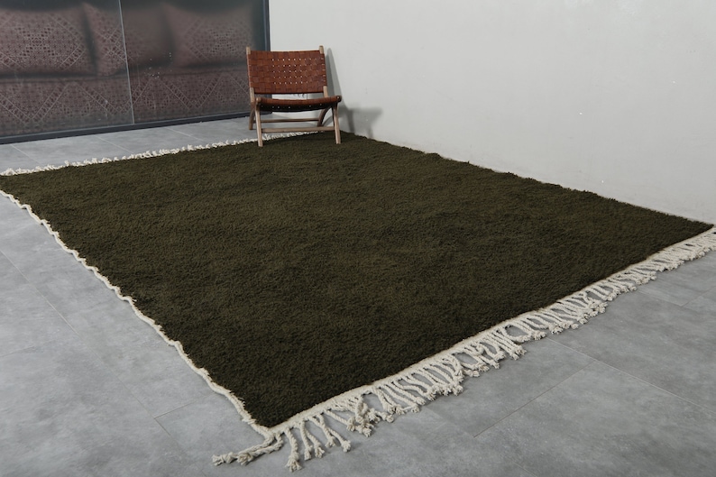 May include: A dark green shag rug with a white fringe border. The rug is laid out on a gray floor.