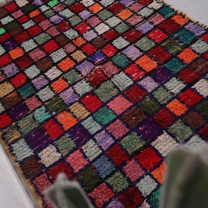 May include: A colorful Moroccan rug with a checkered pattern in shades of red, green, purple, and orange. The rug is made of wool and has a hand-woven texture.