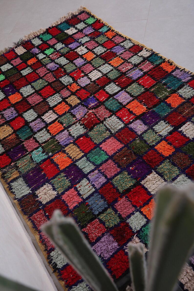 May include: A colorful Moroccan rug with a checkered pattern in shades of red, green, purple, and orange. The rug is made of wool and has a hand-woven texture.