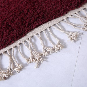 May include: A close-up of a burgundy bath mat with a fringe border. The fringe is made of a natural fiber, like cotton or jute.