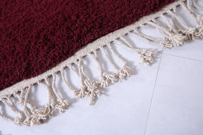 May include: A close-up of a burgundy bath mat with a fringe border. The fringe is made of a natural fiber, like cotton or jute.