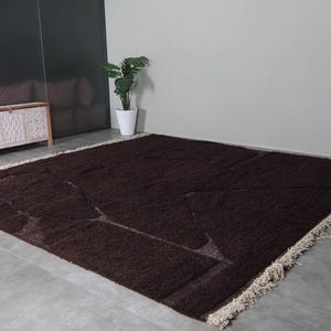 May include: A large, rectangular brown rug with a geometric design. The rug has a shaggy texture and a cream-colored fringe along the edges. The rug is placed on a gray concrete floor.