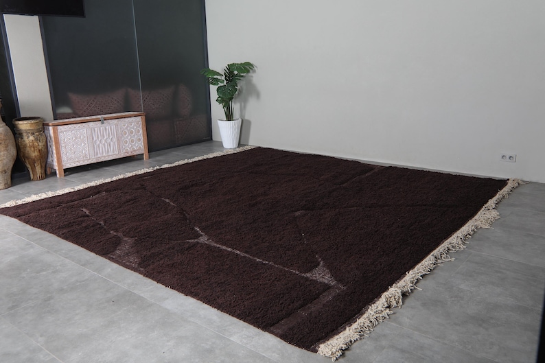 May include: A large, rectangular brown rug with a geometric design. The rug has a shaggy texture and a cream-colored fringe along the edges. The rug is placed on a gray concrete floor.