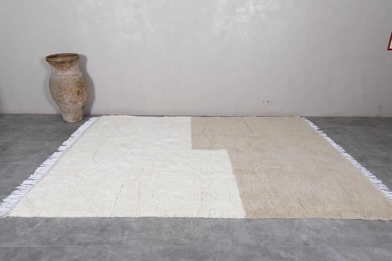 Puede incluir: Una alfombra tejida a mano de color blanco y beige con un dise&ntilde;o geom&eacute;trico. La alfombra tiene un borde de flecos.
