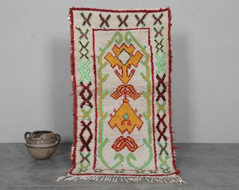 Può includere: Tappeto rettangolare color crema con un disegno centrale arancione, rosso e verde. Il tappeto presenta un motivo geometrico, un bordo rosso e frange sul fondo. Un vaso decorativo in ceramica con manici è a sinistra.