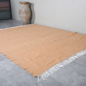 May include: A large, rectangular, peach-colored rug with white fringe around the edges. The rug is lying flat on a gray tiled floor.