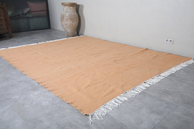 May include: A large, rectangular, peach-colored rug with white fringe around the edges. The rug is lying flat on a gray tiled floor.
