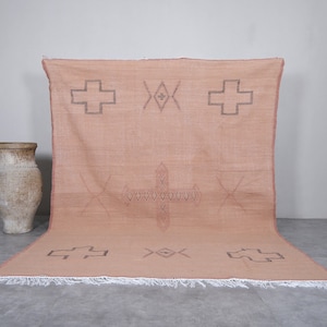 May include: A large, light pink Moroccan rug with a geometric pattern of crosses and diamonds. The rug has a fringe border.