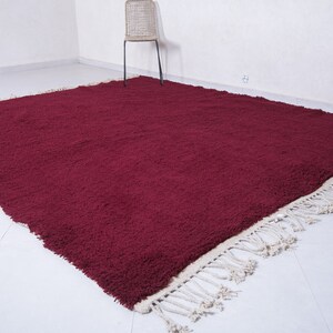 May include: A deep burgundy shag rug with a white fringe border. The rug is laid out on a white tiled floor.