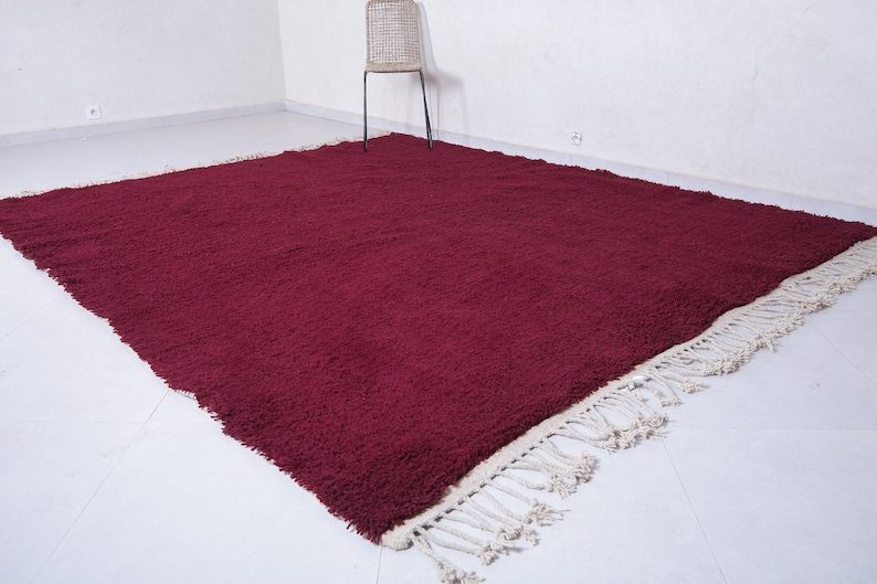 May include: A deep burgundy shag rug with a white fringe border. The rug is laid out on a white tiled floor.
