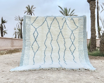 May include: A large, rectangular rug with a cream-colored background and a repeating blue diamond pattern. The rug has a light blue border and fringe along the bottom edge. The rug is displayed outdoors.