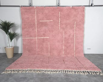 May include: A large, rectangular, pink rug with a geometric design of white lines. The rug has a shaggy texture and a fringe along the bottom edge. The rug is displayed against a white wall.