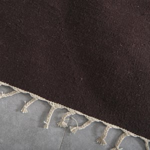 Może przedstawiać: Ciemnobrązowy dywan z frędzlami. Frędzle są wykonane z jasnobeżowego materiału.
