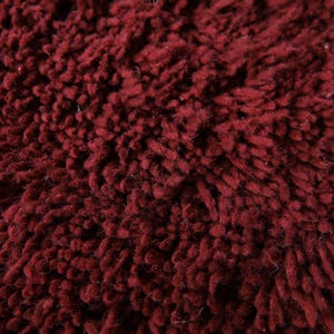 May include: Close-up of a deep burgundy, textured fabric. The material has a thick, looped pile, creating a plush, soft appearance. The color is a rich, dark red, with subtle variations in tone across the surface.