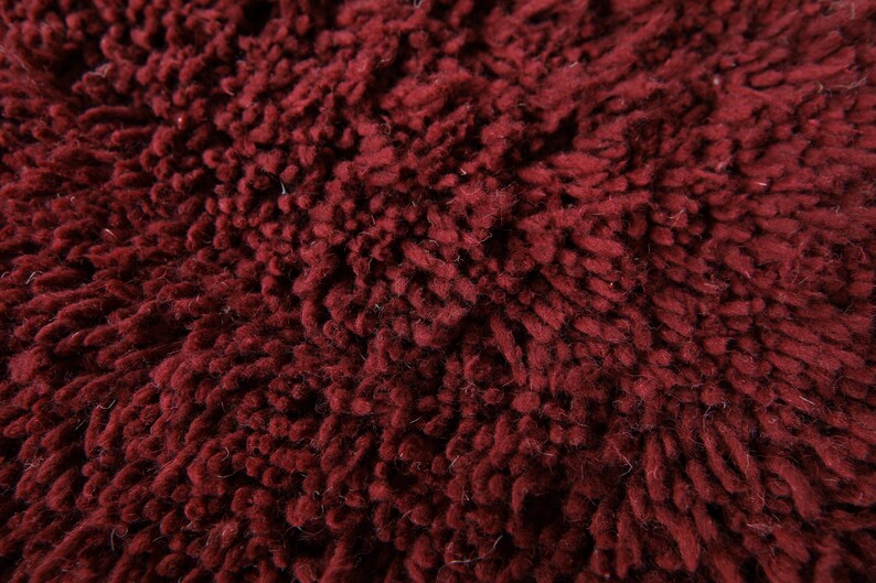 May include: Close-up of a deep burgundy, textured fabric. The material has a thick, looped pile, creating a plush, soft appearance. The color is a rich, dark red, with subtle variations in tone across the surface.