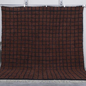 Peut inclure: Un grand tapis rectangulaire marron avec un motif de grille noire. Le tapis a une texture touffue et une frange blanche le long du bord inf&eacute;rieur. Le tapis est pr&eacute;sent&eacute; contre un mur blanc, avec un vase d&eacute;coratif sur la gauche.