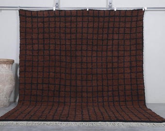 May include: A large, rectangular brown rug with a black grid pattern. The rug has a shaggy texture and a white fringe along the bottom edge. The rug is displayed against a white wall, with a decorative vase on the left.