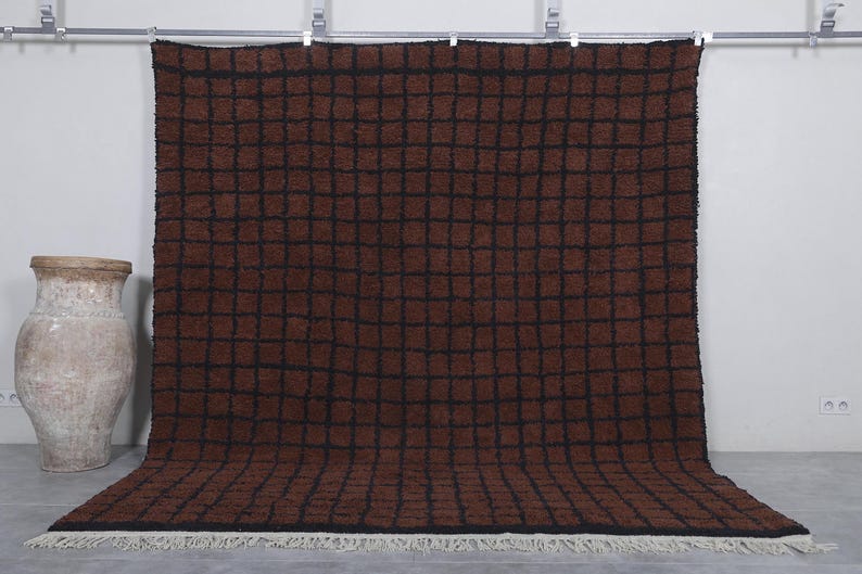 Peut inclure: Un grand tapis rectangulaire marron avec un motif de grille noire. Le tapis a une texture touffue et une frange blanche le long du bord inf&eacute;rieur. Le tapis est pr&eacute;sent&eacute; contre un mur blanc, avec un vase d&eacute;coratif sur la gauche.