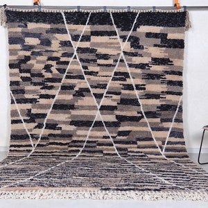 May include: A large, hand-woven rug with a geometric pattern in shades of beige, black, and white. The rug has a fringe border and is hanging on a wall.