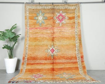 May include: A large, rectangular rug with a warm color palette of orange, yellow, and pink. The rug features geometric patterns in the center and along the border, with a white and cream-colored border. The rug is displayed against a neutral wall.