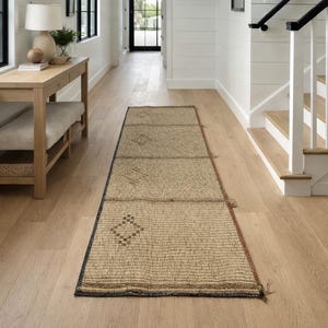 May include: A rectangular, woven runner rug with a geometric pattern in shades of tan and brown. The rug is placed on a light wood floor in a hallway. The rug has a dark border and is made of natural fibers.