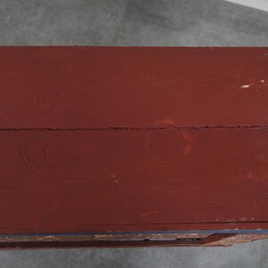 May include: A close-up of a wooden chest with a deep red painted surface. The chest's top is made of planks, showing the wood grain. The edges are trimmed with a darker color, and the chest appears aged.
