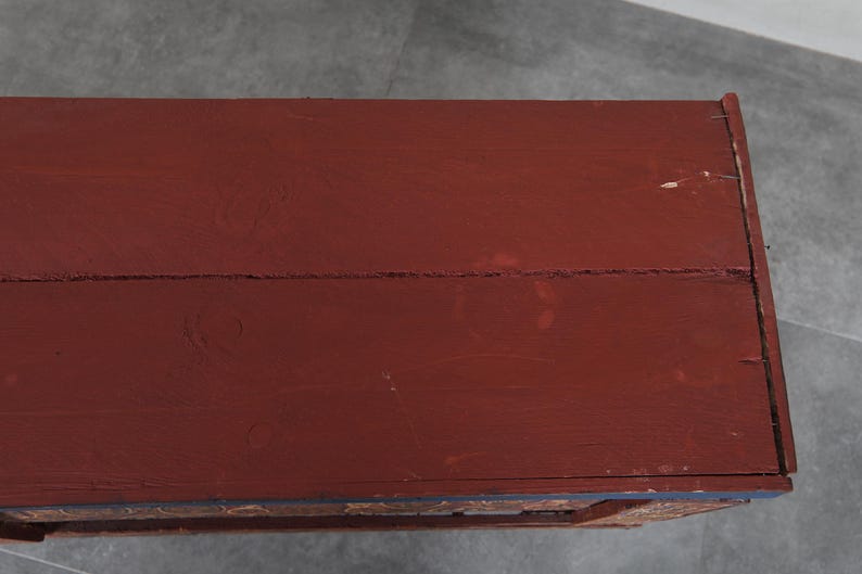 May include: A close-up of a wooden chest with a deep red painted surface. The chest's top is made of planks, showing the wood grain. The edges are trimmed with a darker color, and the chest appears aged.