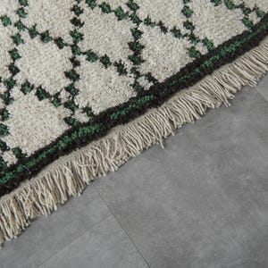 K&ouml;nnte beinhalten: Nahaufnahme eines cremefarbenen Teppichs mit einem geometrischen Muster in Dunkelgr&uuml;n und Schwarz. Der Teppich hat einen Fransenrand. Der Teppich liegt auf einer grauen Oberfl&auml;che.