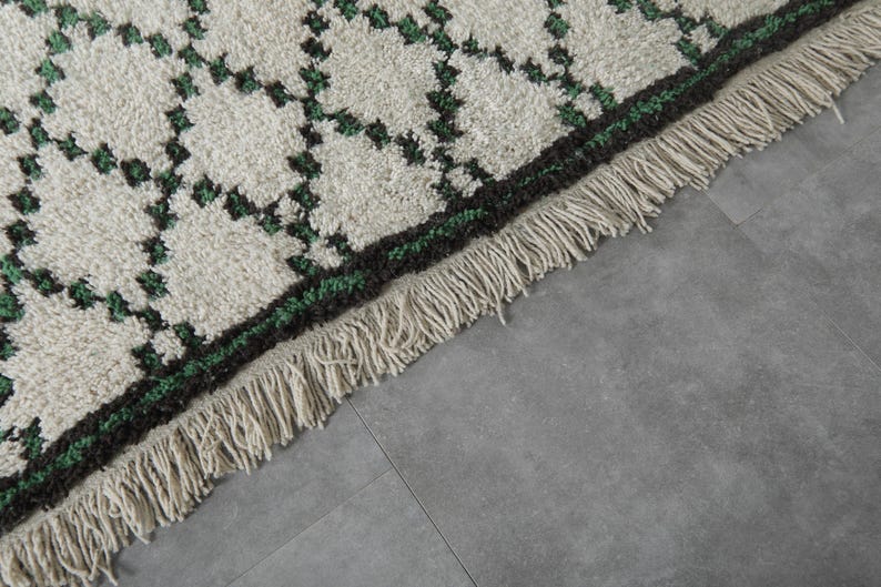 K&ouml;nnte beinhalten: Nahaufnahme eines cremefarbenen Teppichs mit einem geometrischen Muster in Dunkelgr&uuml;n und Schwarz. Der Teppich hat einen Fransenrand. Der Teppich liegt auf einer grauen Oberfl&auml;che.