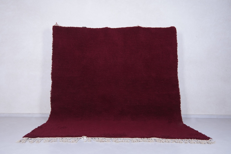 May include: A deep burgundy shag rug with a white fringe border. The rug is folded in half and lying on a white surface.