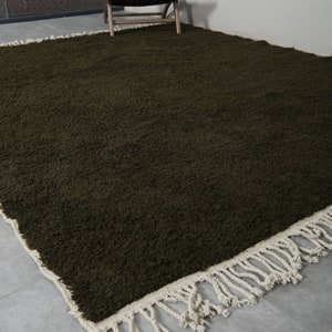 May include: A dark green shag rug with a white fringe border. The rug is on a gray tiled floor and a wooden chair is in the background.