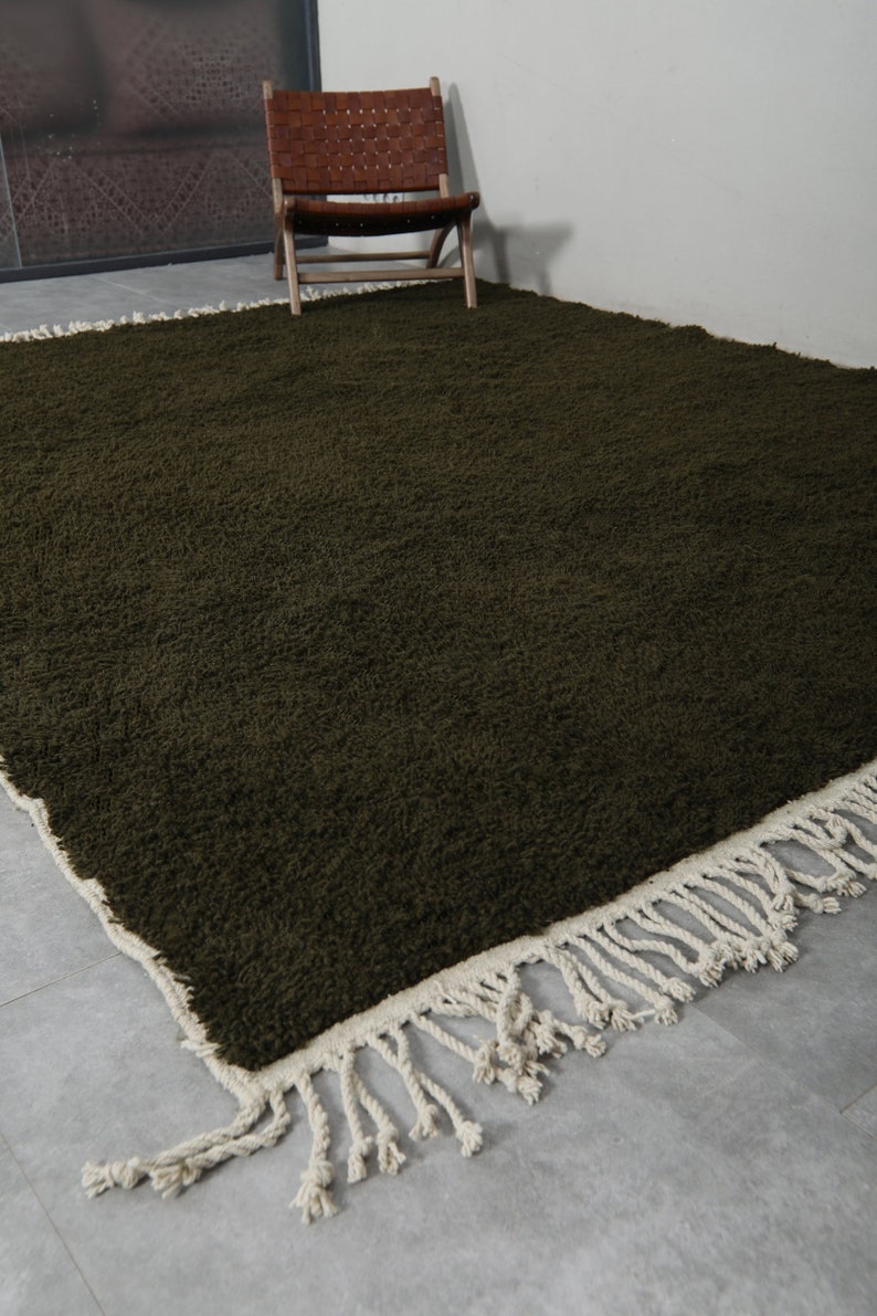 May include: A dark green shag rug with a white fringe border. The rug is on a gray tiled floor and a wooden chair is in the background.