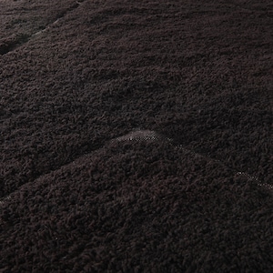 May include: Close-up of a dark brown, textured fabric. The material appears to be plush and soft, with a slightly uneven surface. The fabric is folded, creating diagonal lines across the image.