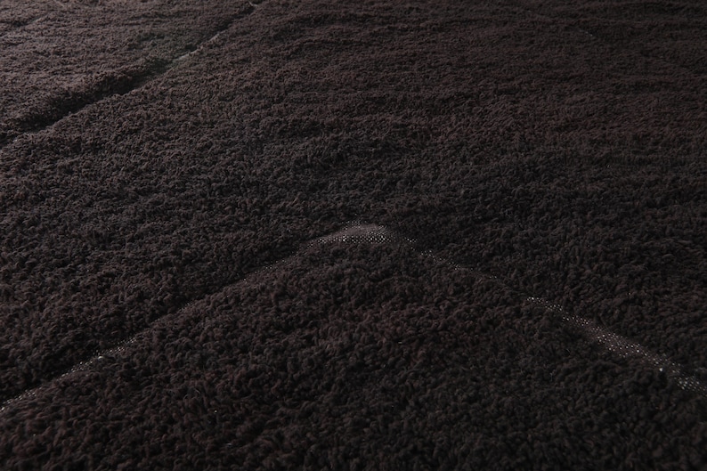 May include: Close-up of a dark brown, textured fabric. The material appears to be plush and soft, with a slightly uneven surface. The fabric is folded, creating diagonal lines across the image.