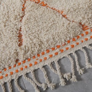 May include: Close-up of a cream-colored, textured rug with an orange geometric design. The rug has a fringe border with knotted tassels. The design features orange stitching and a soft, plush texture, suitable for home decor.