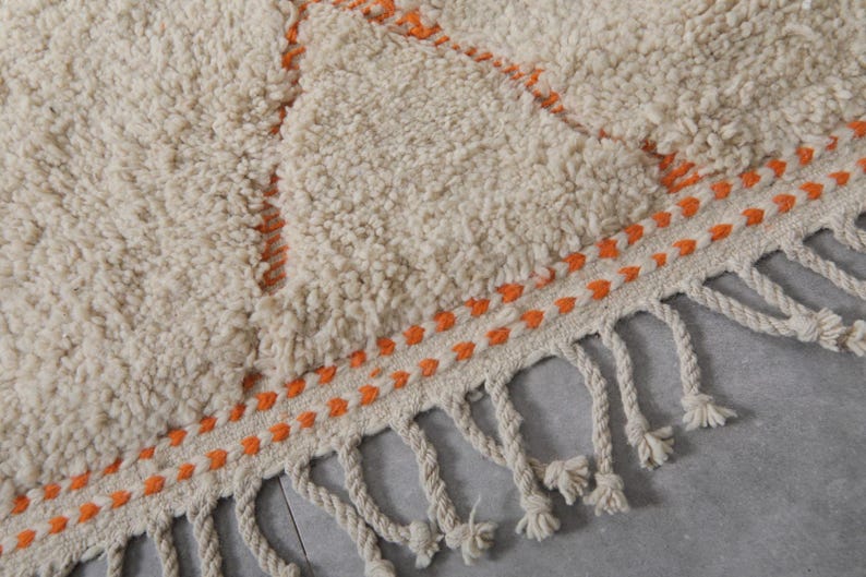 May include: Close-up of a cream-colored, textured rug with an orange geometric design. The rug has a fringe border with knotted tassels. The design features orange stitching and a soft, plush texture, suitable for home decor.