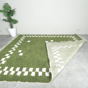 May include: Green and white geometric patterned rug. The rug has a shaggy texture and a rectangular shape. The design features a border of white squares and a matching pattern in the center. The rug is displayed on a gray floor.