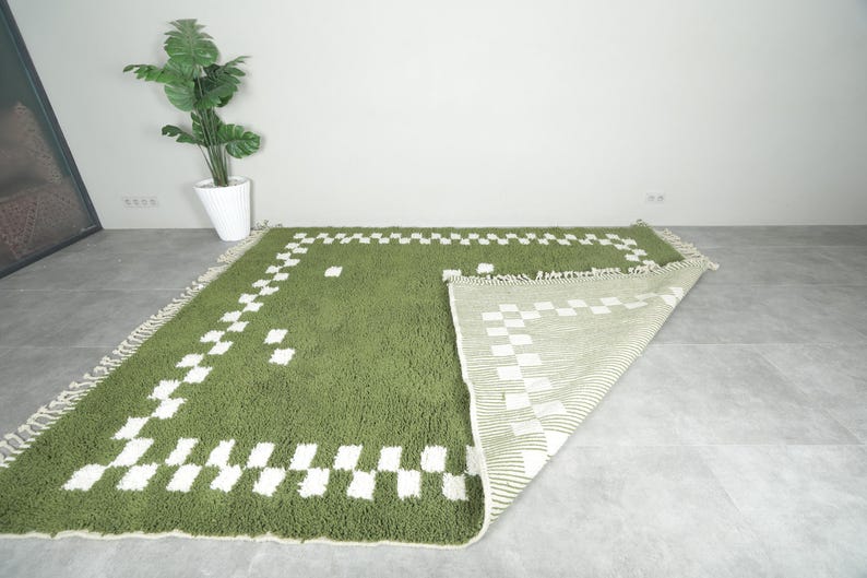 May include: Green and white geometric patterned rug. The rug has a shaggy texture and a rectangular shape. The design features a border of white squares and a matching pattern in the center. The rug is displayed on a gray floor.