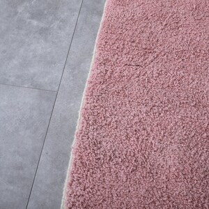 May include: Close-up of a soft, pink, fluffy rug with a white border. The rug is placed on a gray tiled floor.